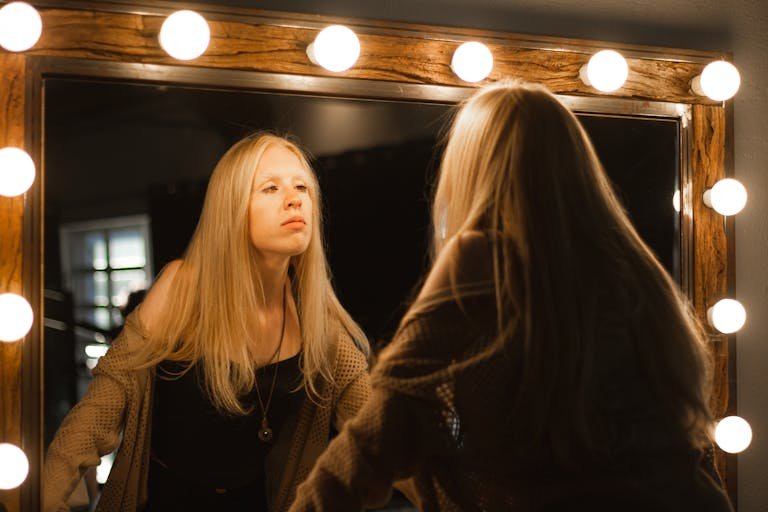 A blonde woman gazes at her reflection in a warmly lit vanity mirror, creating a serene and introspective mood.