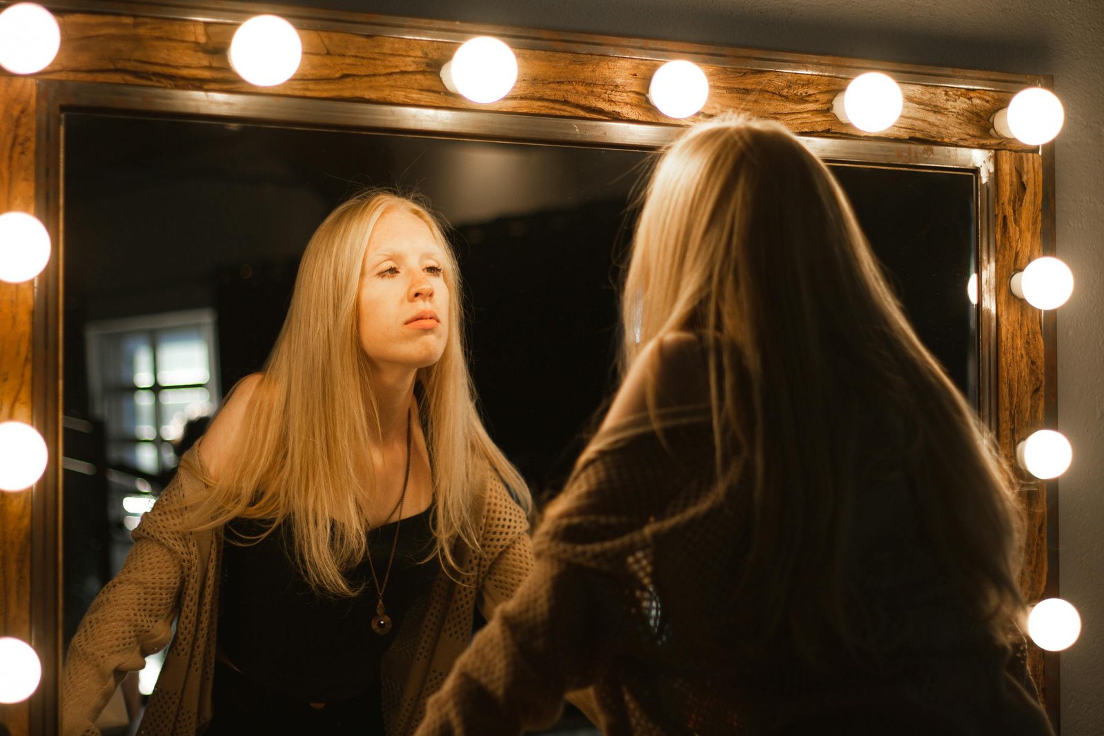 A blonde woman gazes at her reflection in a warmly lit vanity mirror, creating a serene and introspective mood.