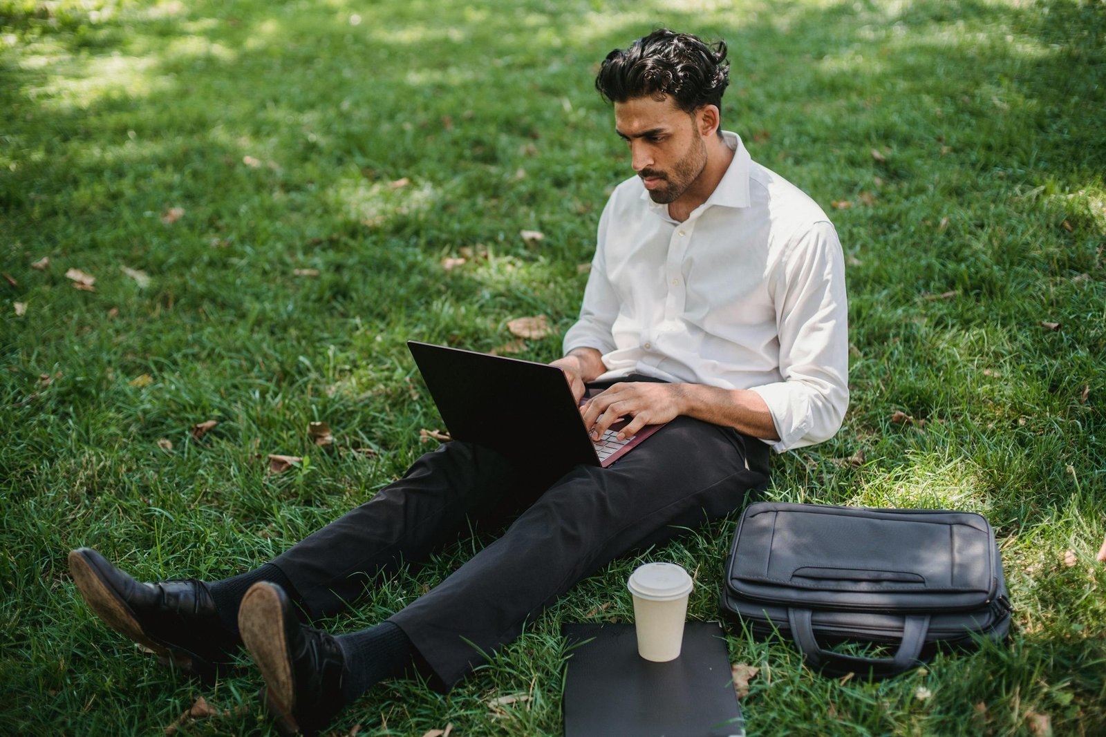 A man in a white shirt working remotely on his laptop in a park, embracing modern technology.
