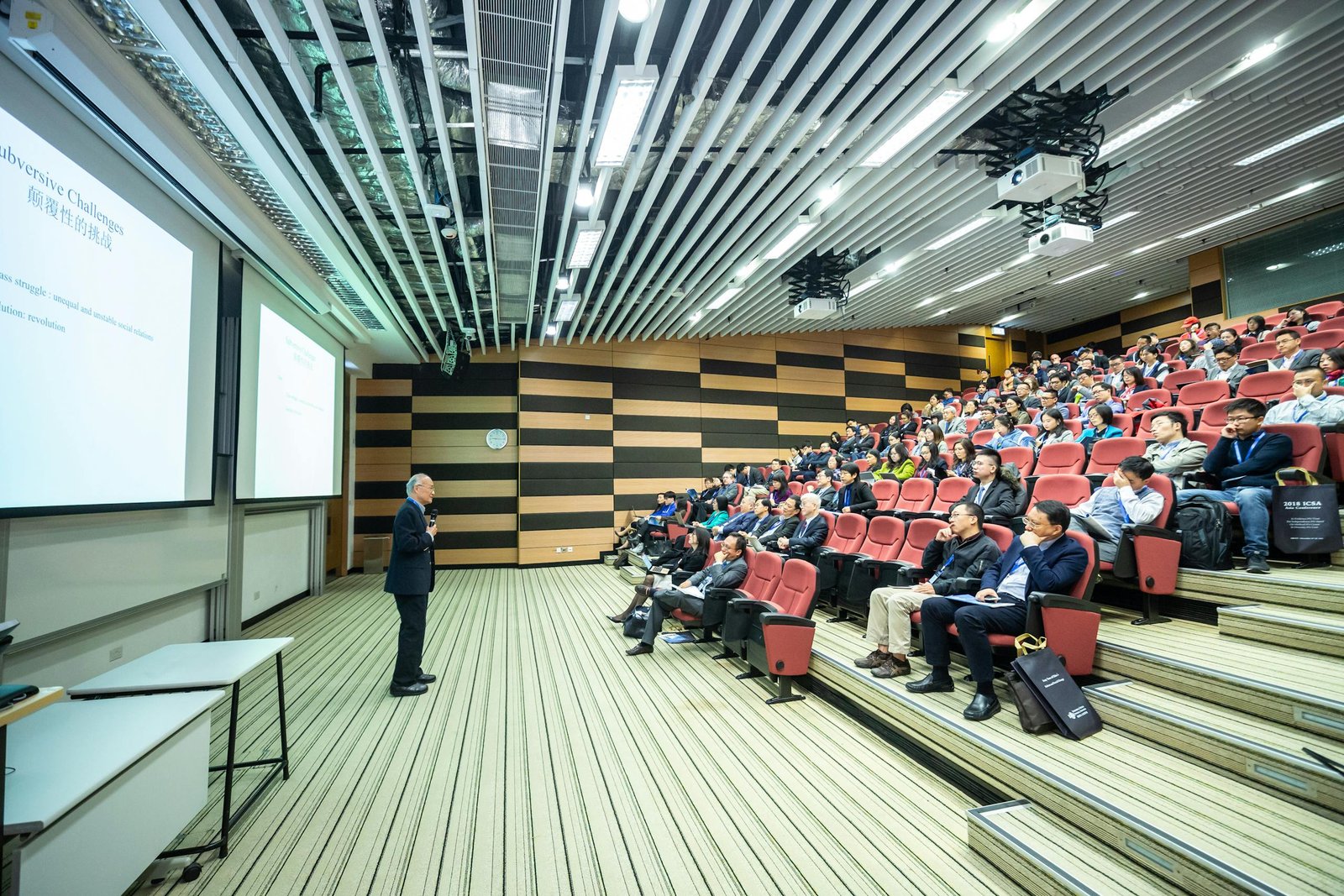 A speaker delivers a presentation to an attentive audience in a modern auditorium setting.