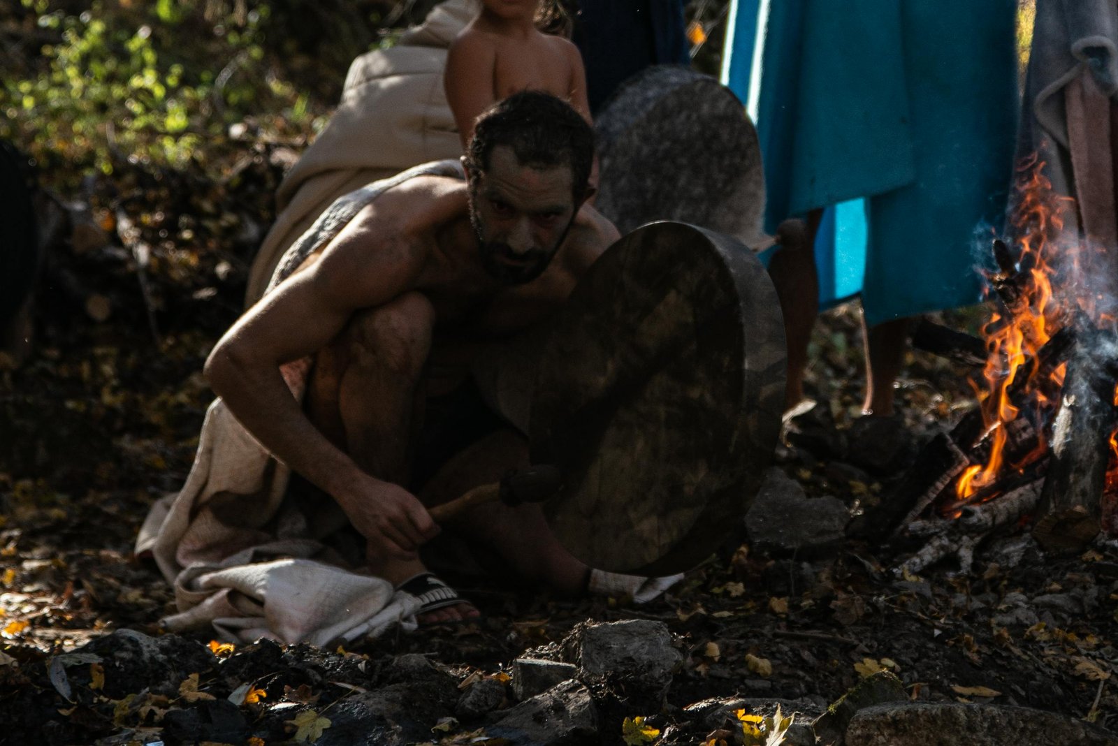 Man playing drum by campfire surrounded by nature and people.