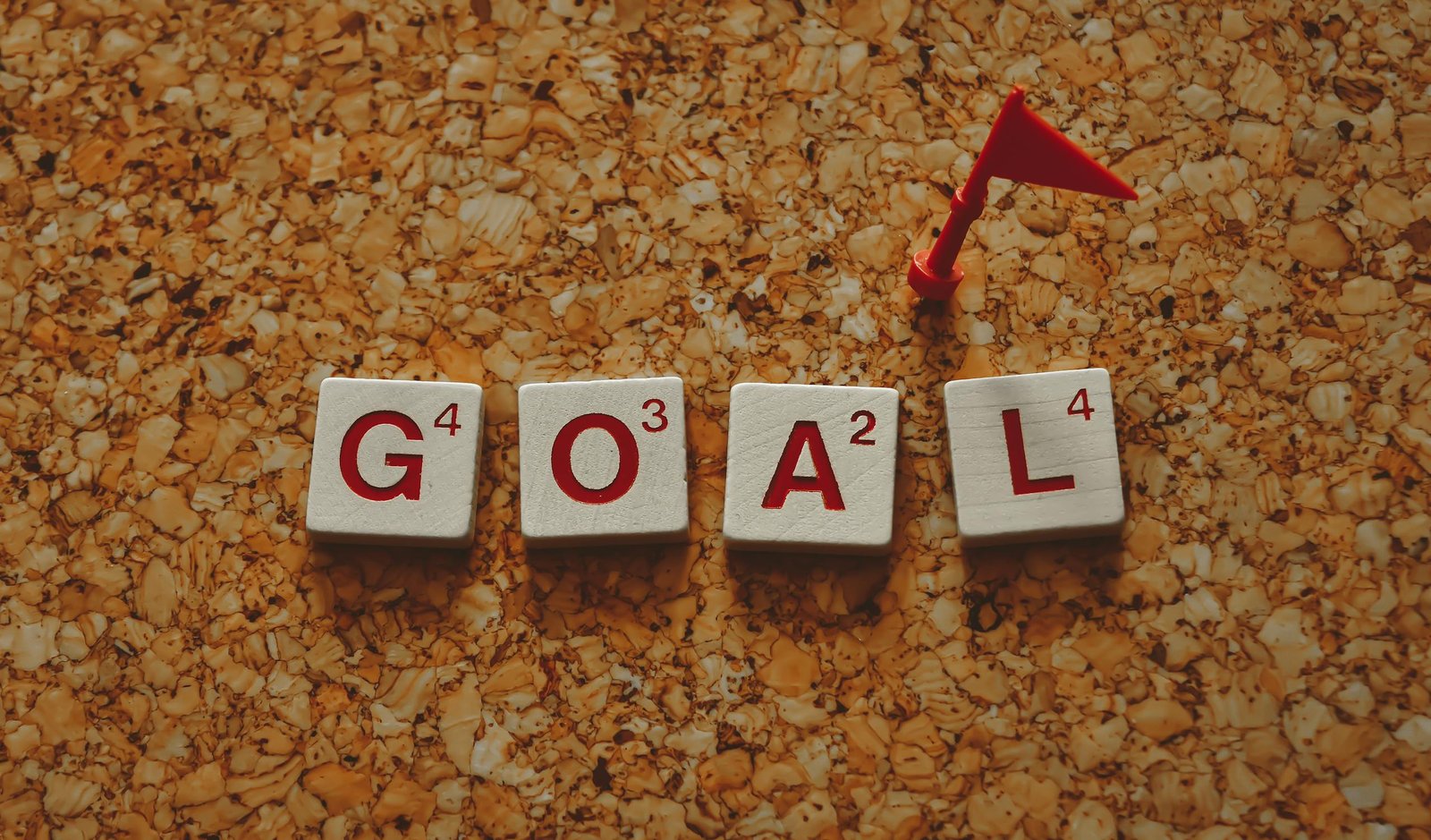 Scrabble tiles forming the word 'goal' with a small red flag on a cork board background.